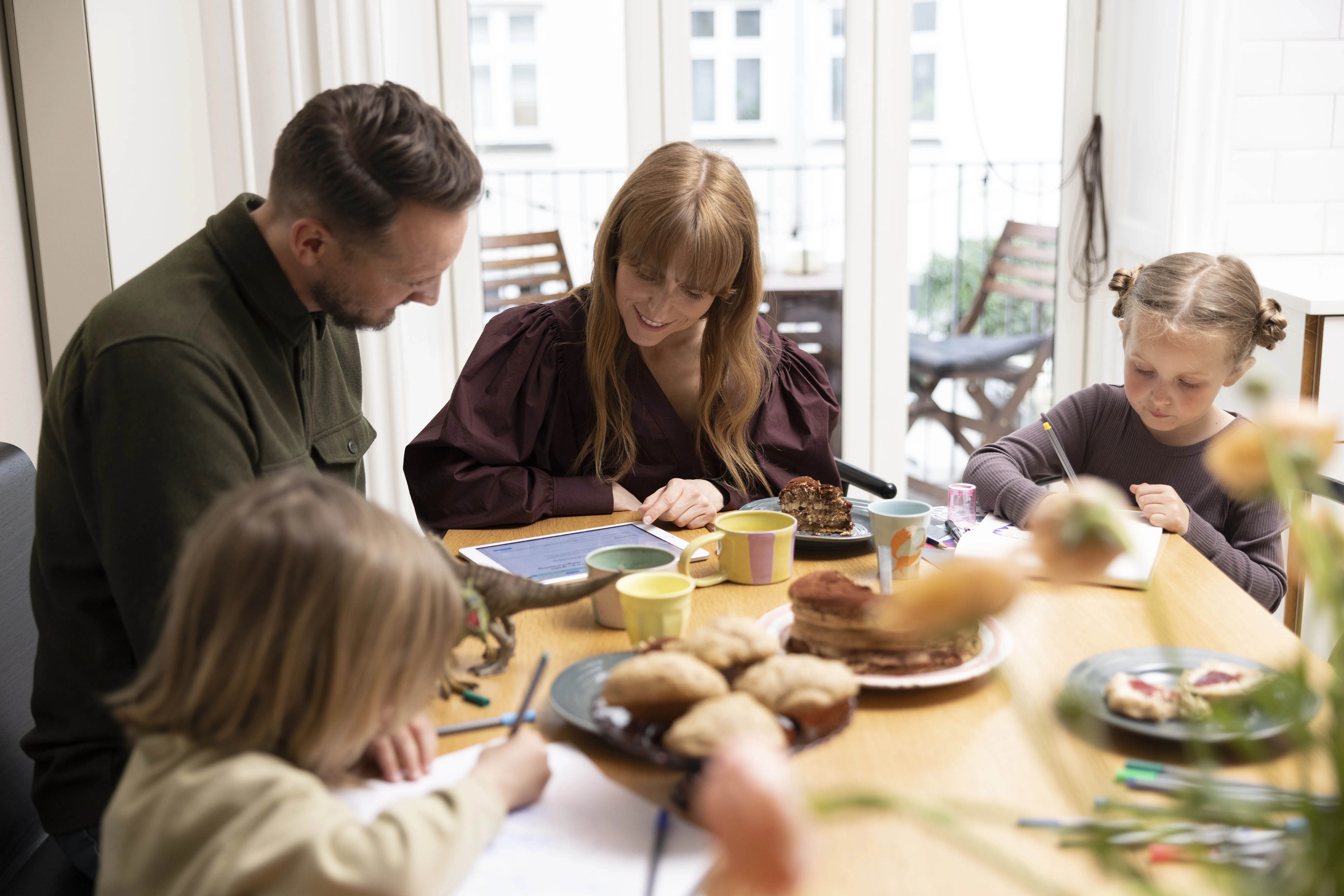 Familie med to børn ved morgenbord, voksne ser på tablet mens børnene tegner