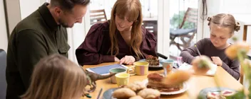 Familie med to børn ved morgenbord, voksne ser på tablet mens børnene tegner