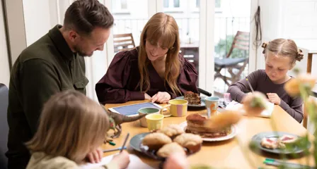 Familie med to børn ved morgenbord, voksne ser på tablet mens børnene tegner