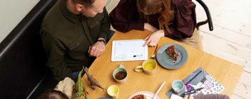 Familie med to børn ved bord, voksne ser på tablet, børn tegner