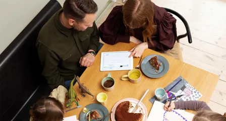 Familie med to børn ved bord, voksne ser på tablet, børn tegner