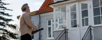 Ejendomsmægler foran hus med notesblok laver en vurdering af bolig med altan og hvide vinduer