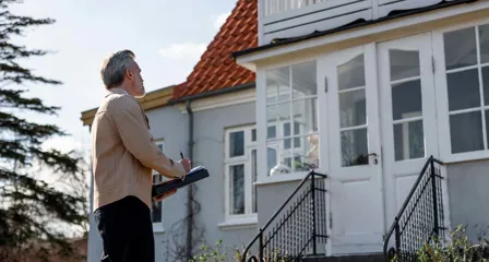 Ejendomsmægler foran hus med notesblok laver en vurdering af bolig med altan og hvide vinduer