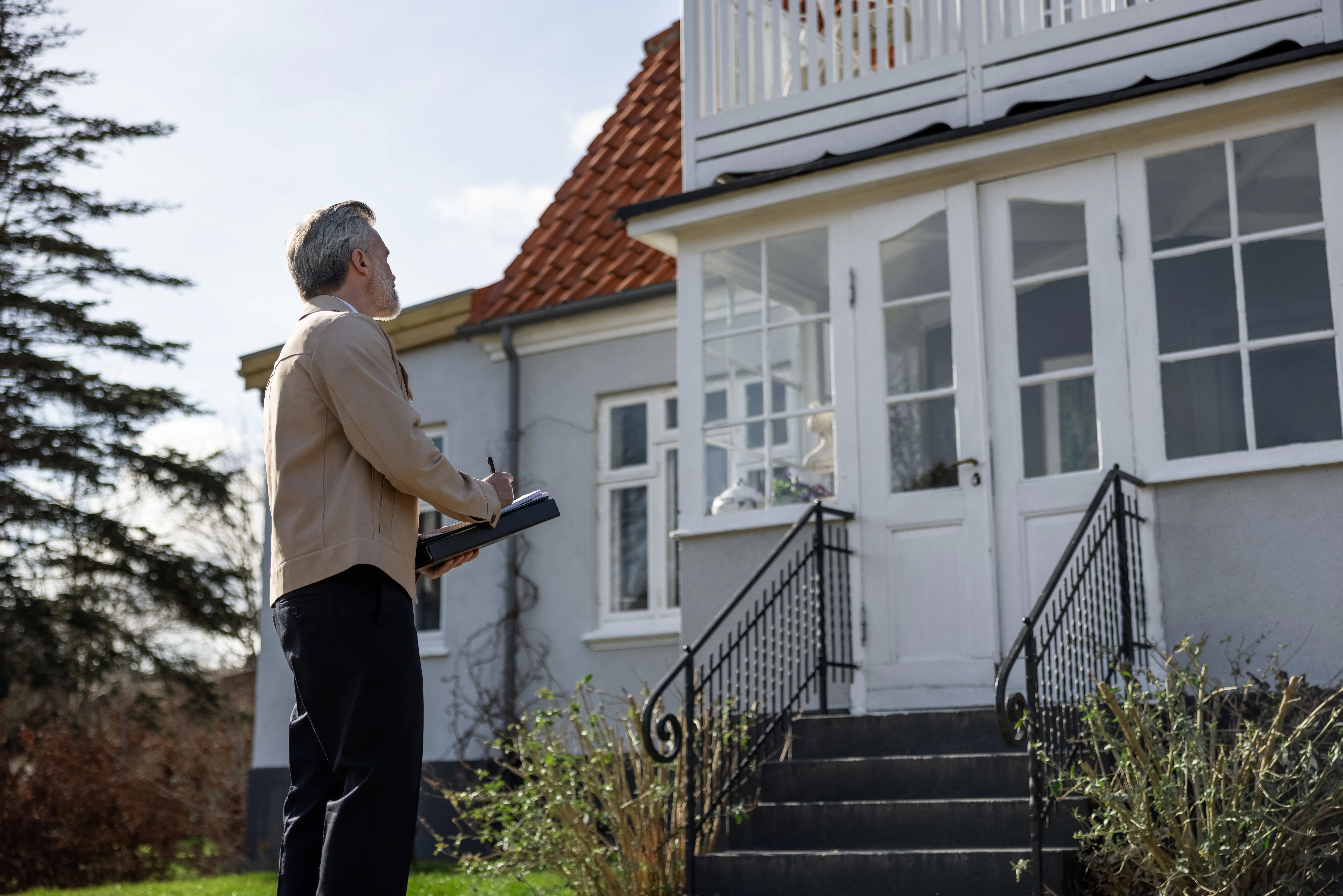 Ejendomsmægler foran hus med notesblok laver en vurdering af bolig med altan og hvide vinduer