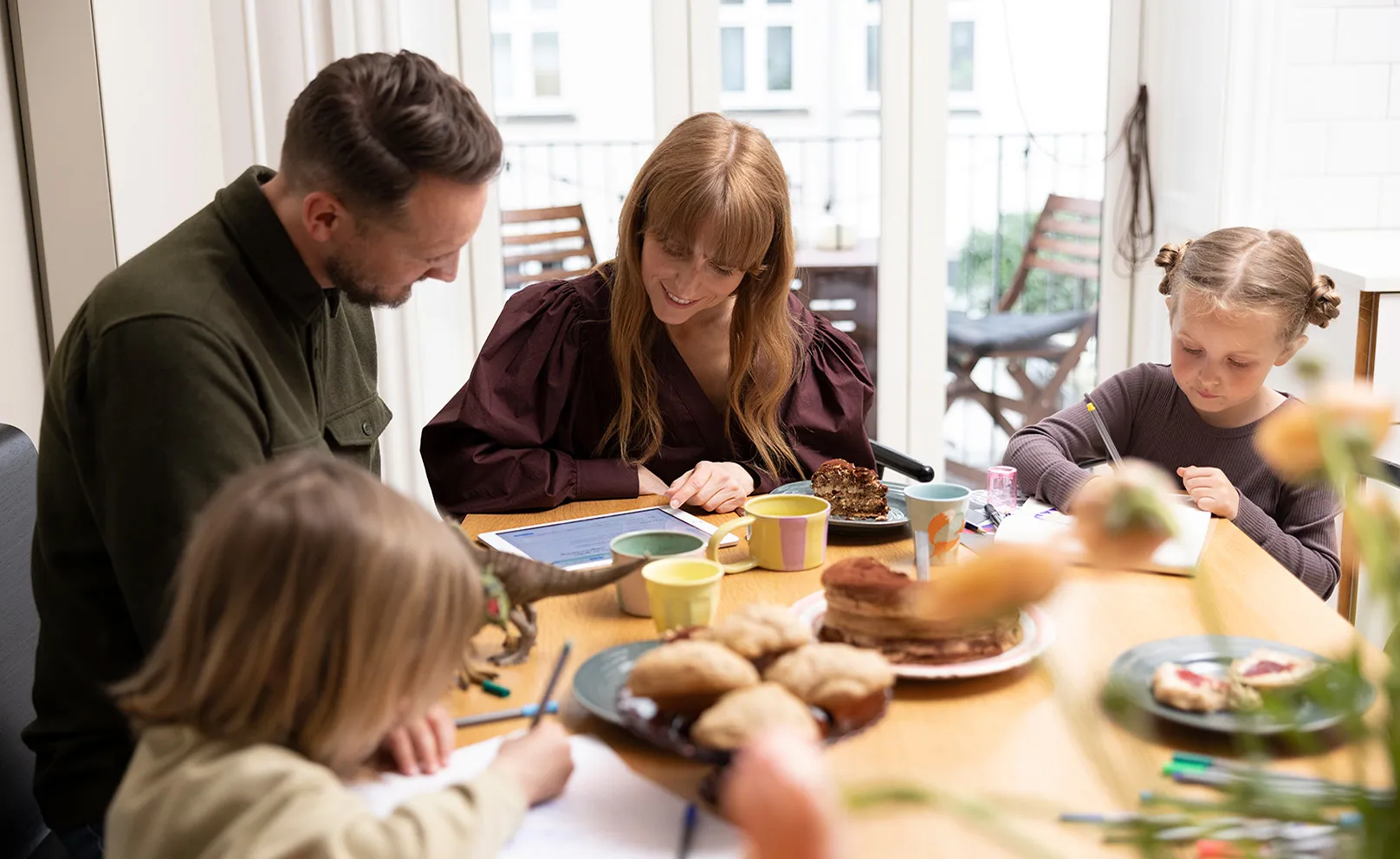 Familie med to børn ved morgenbord, voksne ser på tablet mens børnene tegner
