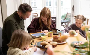 Familie med to børn ved morgenbord, voksne ser på tablet mens børnene tegner